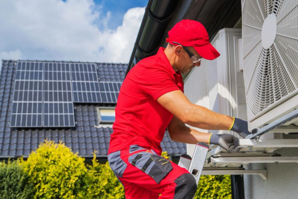 A technician in a red cap and shirt services an outdoor air conditioning unit on a ladder.