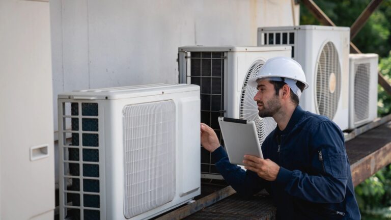 A male technician in a hard hat inspects an outdoor AC unit while holding a tablet.