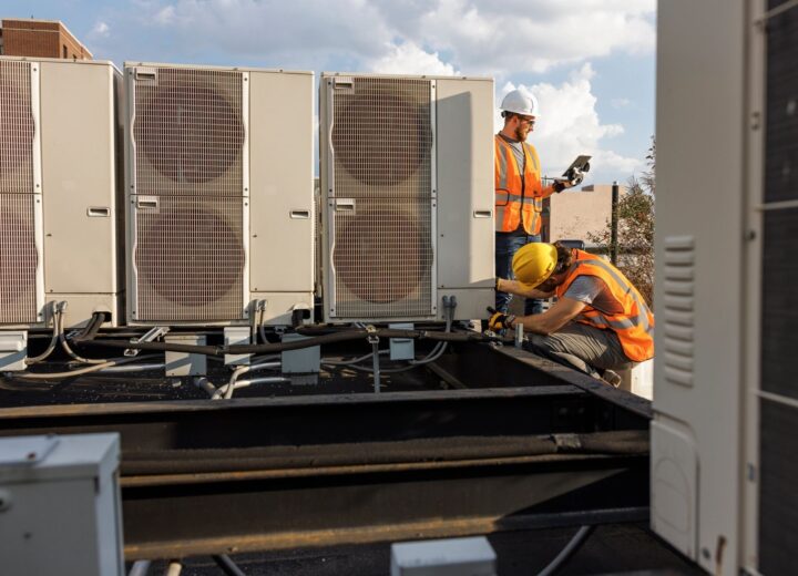 Two HVAC technicians in safety vests and hard hats work on commercial rooftop units.