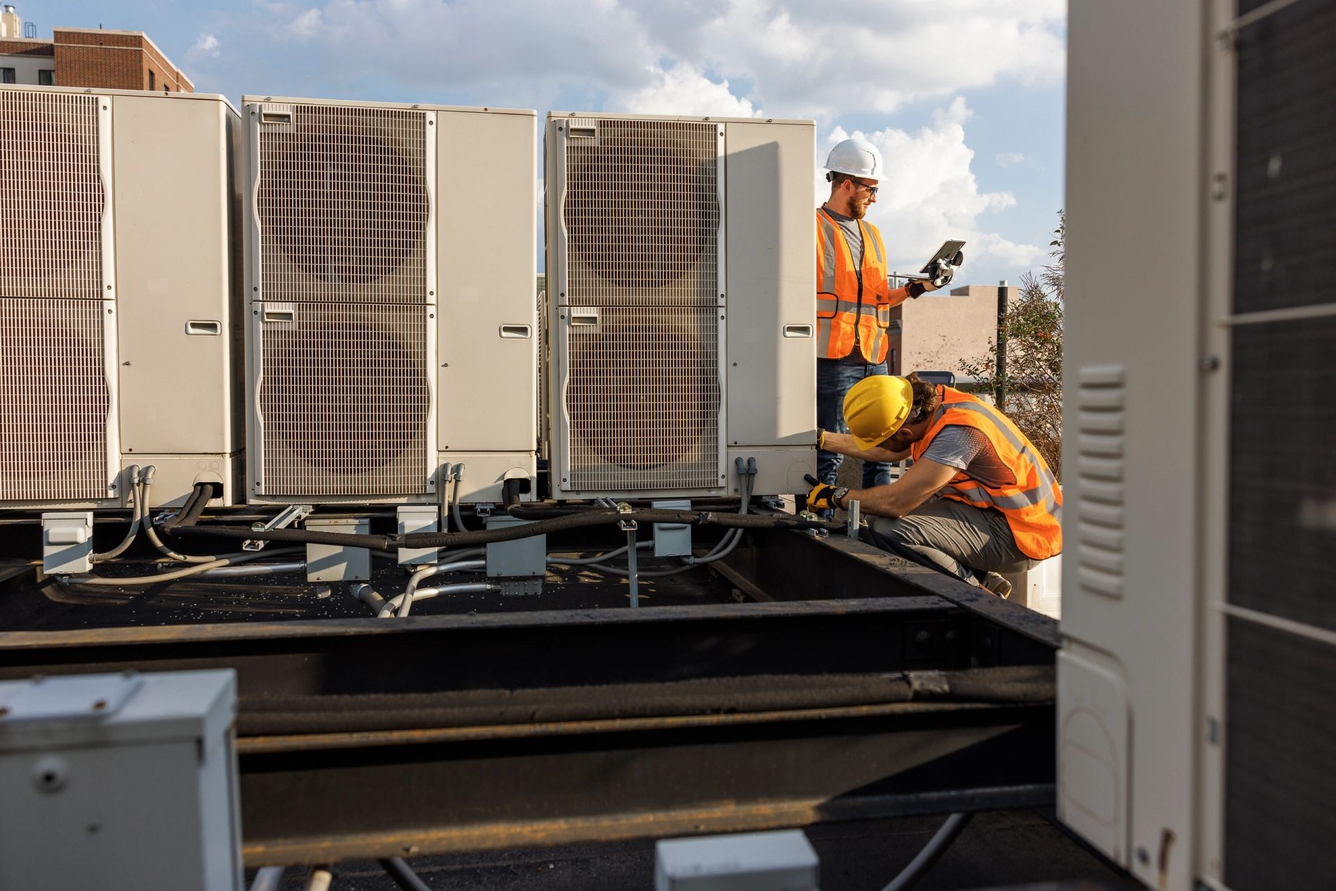 Two HVAC technicians in safety vests and hard hats work on commercial rooftop units.