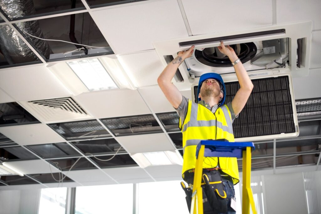 A technician in a hard hat and safety vest installs a ceiling air conditioning unit from a ladder.