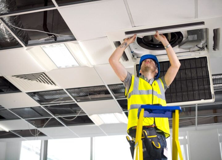 A technician in a hard hat and safety vest installs a ceiling air conditioning unit from a ladder.