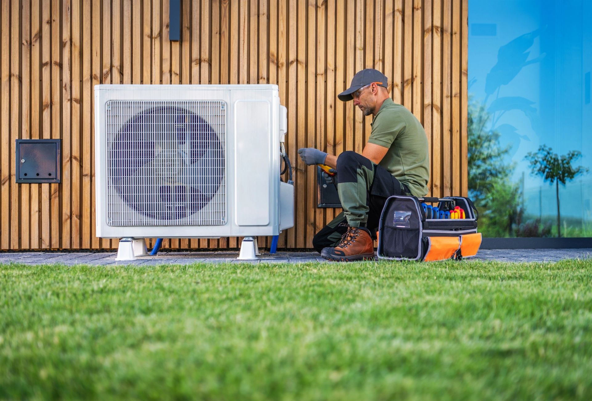 A male technician in a cap and gloves services an outdoor HVAC unit next to a wooden wall.