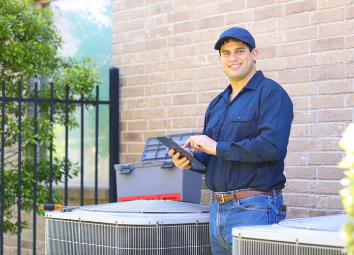 A smiling male technician in a cap stands next to an outdoor HVAC unit, holding a tablet.