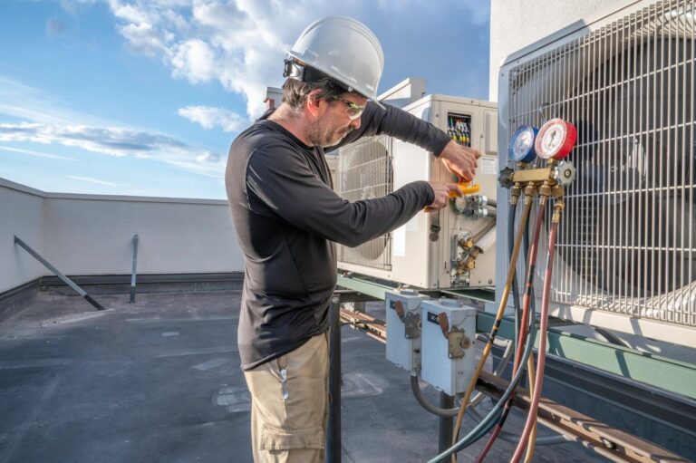 A male technician in a hard hat and safety glasses services a rooftop AC unit with gauges.